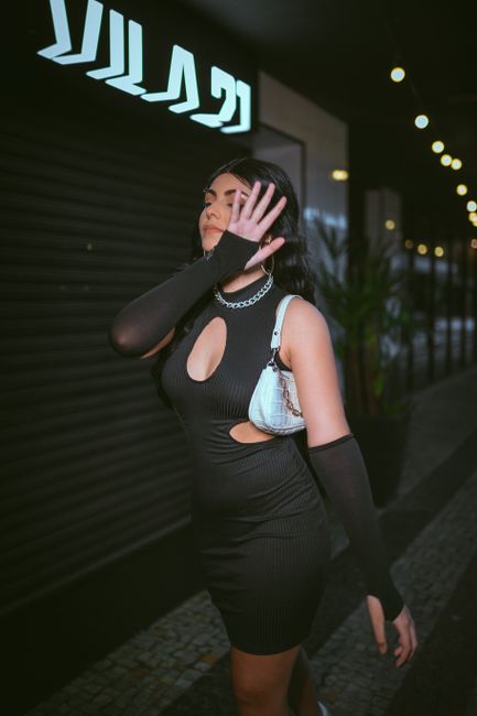 Woman in a black dress poses with hand covering face in an urban night scene.