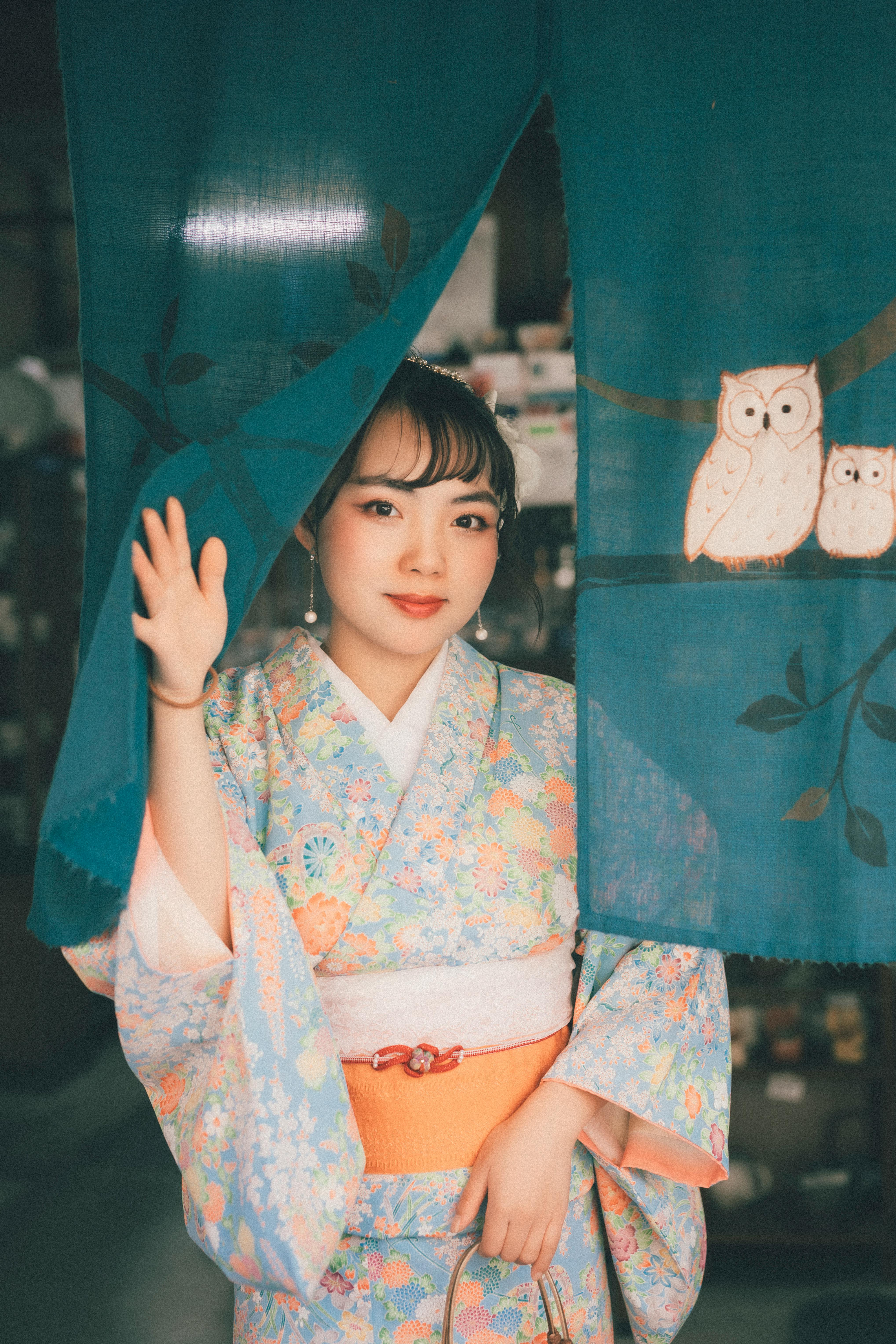 A young woman in a floral kimono opens a traditional Japanese curtain with owl designs.