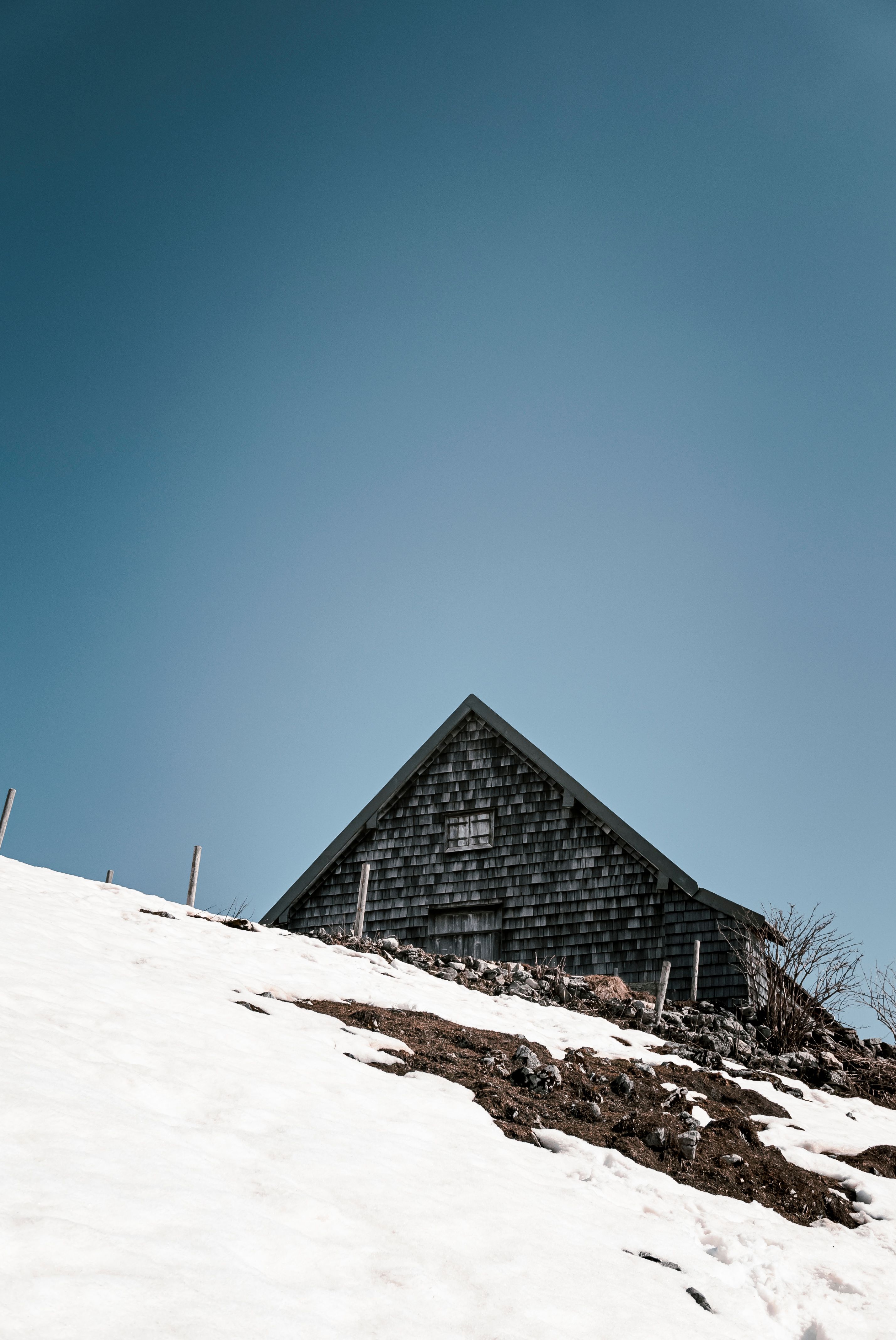 A wooden cabin rests on a snow-covered hill beneath a clear blue sky, creating a serene rural winter scene.