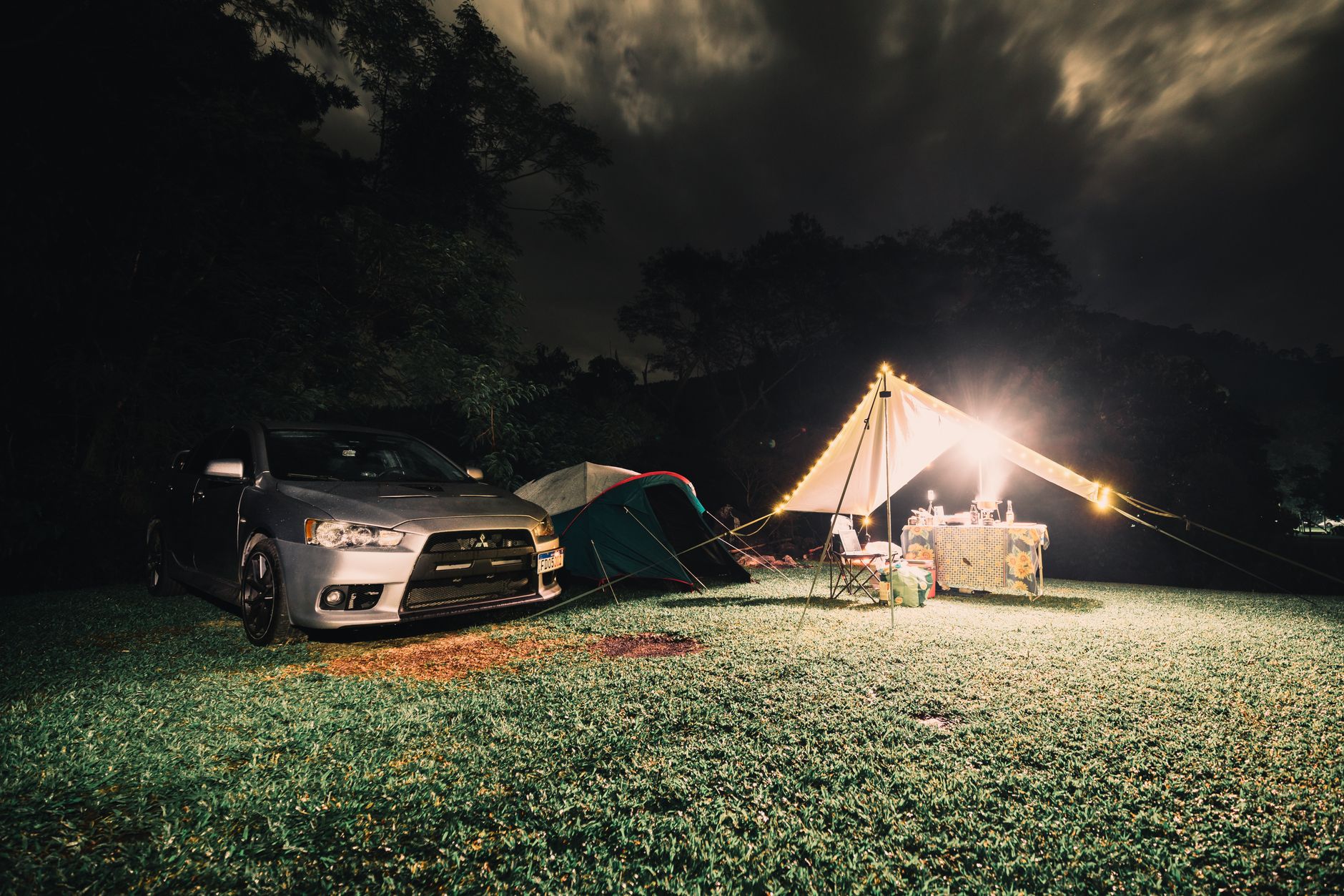 Serene camping site at night featuring illuminated tents, car, and lush grass under a cloudy sky.