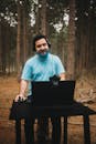 Smiling Man in Headphones Sitting at Laptop Desk in Forest