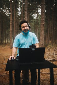 Man smiling while working on a laptop outdoors in a forest setting, enjoying remote work.