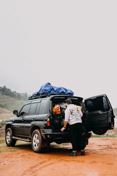 A man packs an SUV with luggage on a dirt road, preparing for an outdoor adventure.