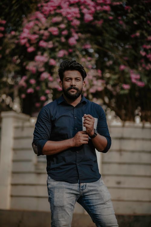 Free A confident man stands outdoors with a floral backdrop, exuding casual charm and style. Stock Photo