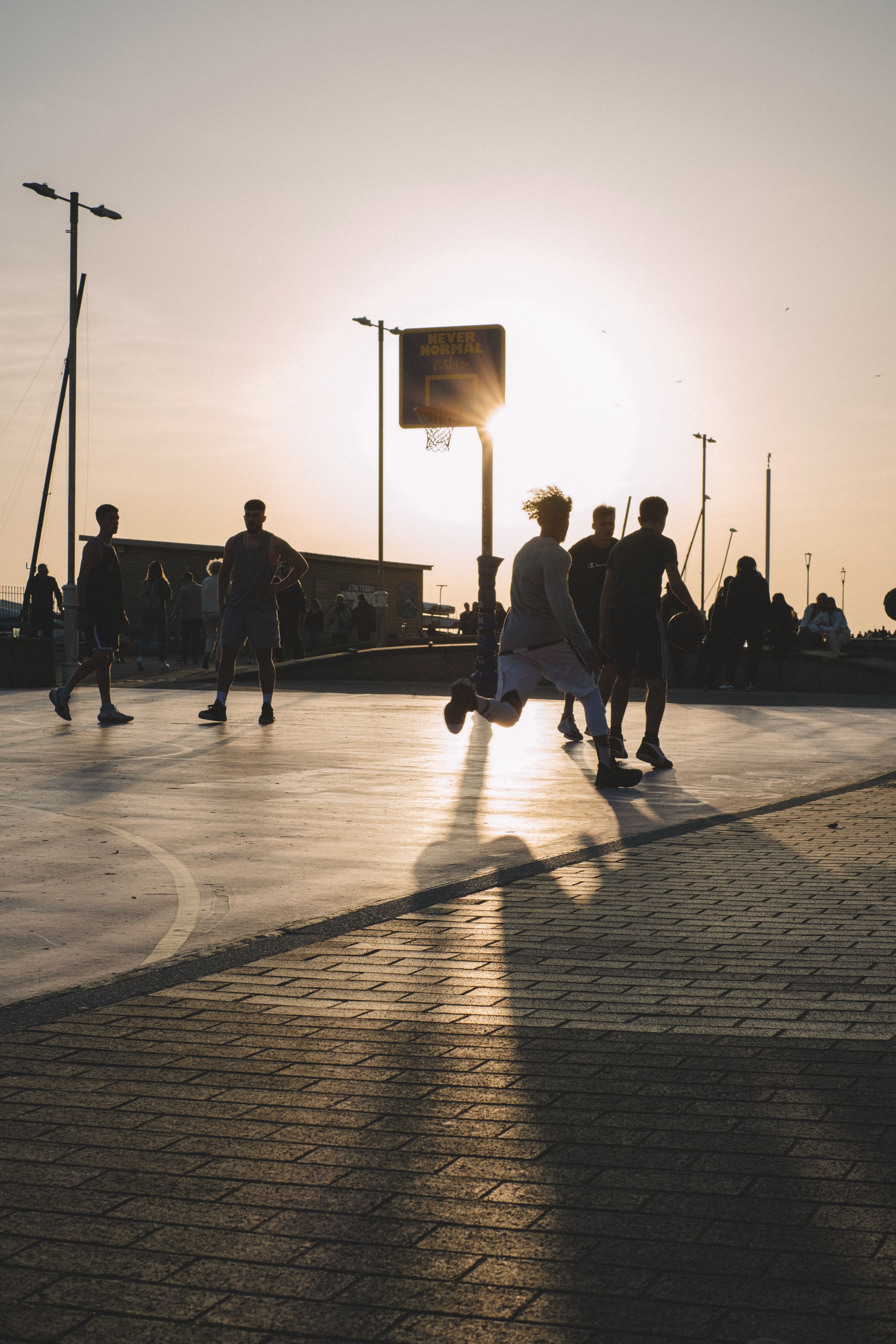 Silhouettes of people playing basketball on an outdoor court at sunset with long shadows.