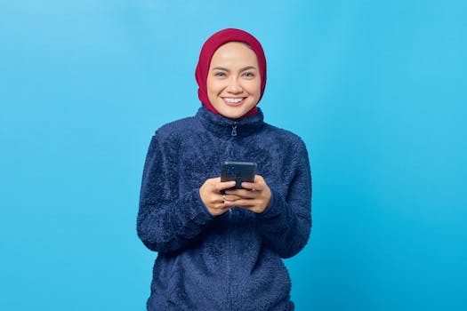 Smiling young woman in hijab wearing a fur jacket, using smartphone against a blue background.