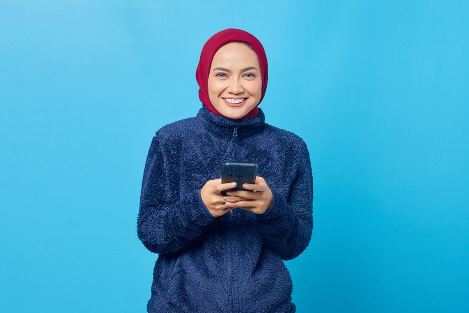 Smiling young woman in hijab wearing a fur jacket, using smartphone against a blue background.