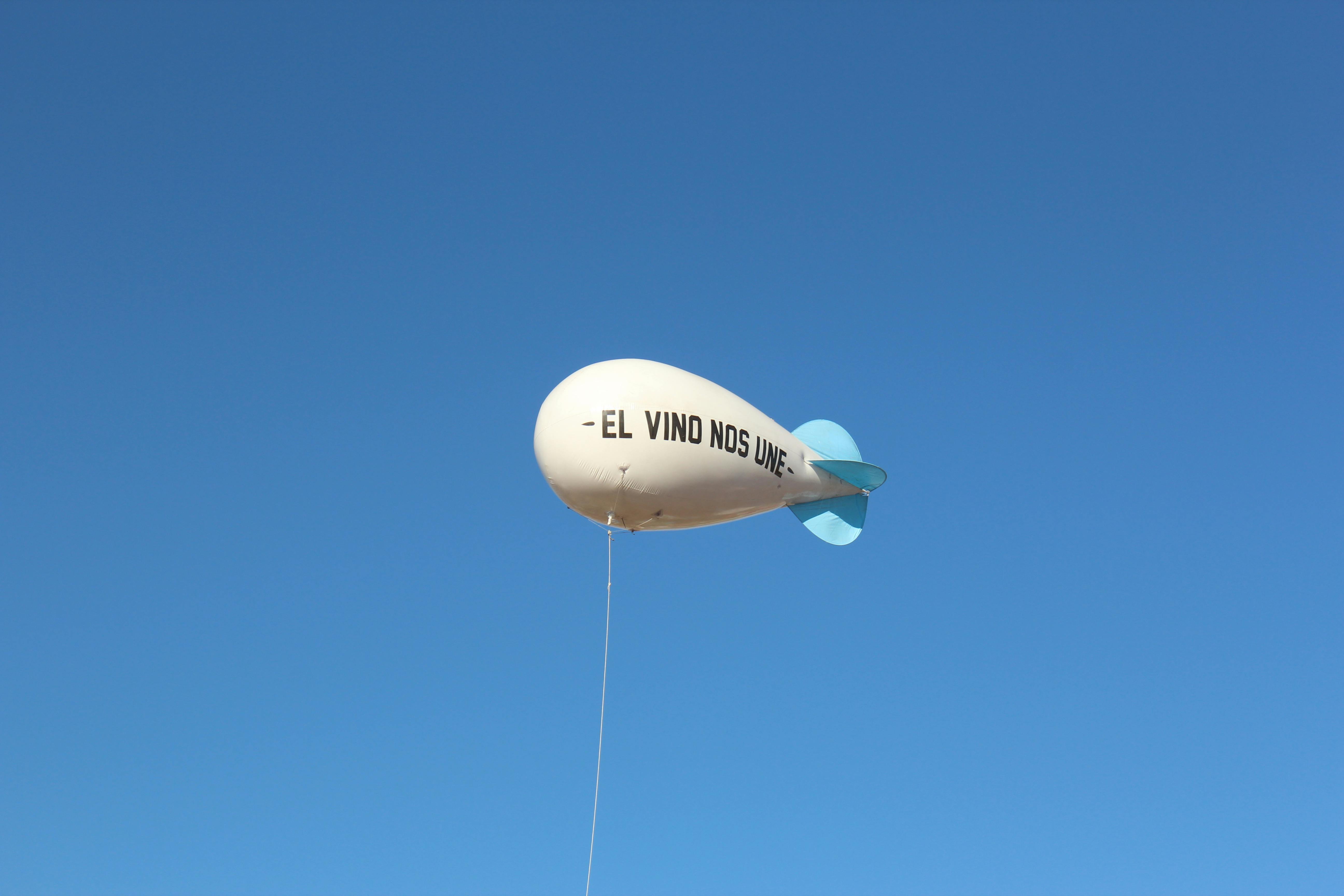 A white balloon with 'El Vino Nos Une' phrase floats against a clear blue sky.