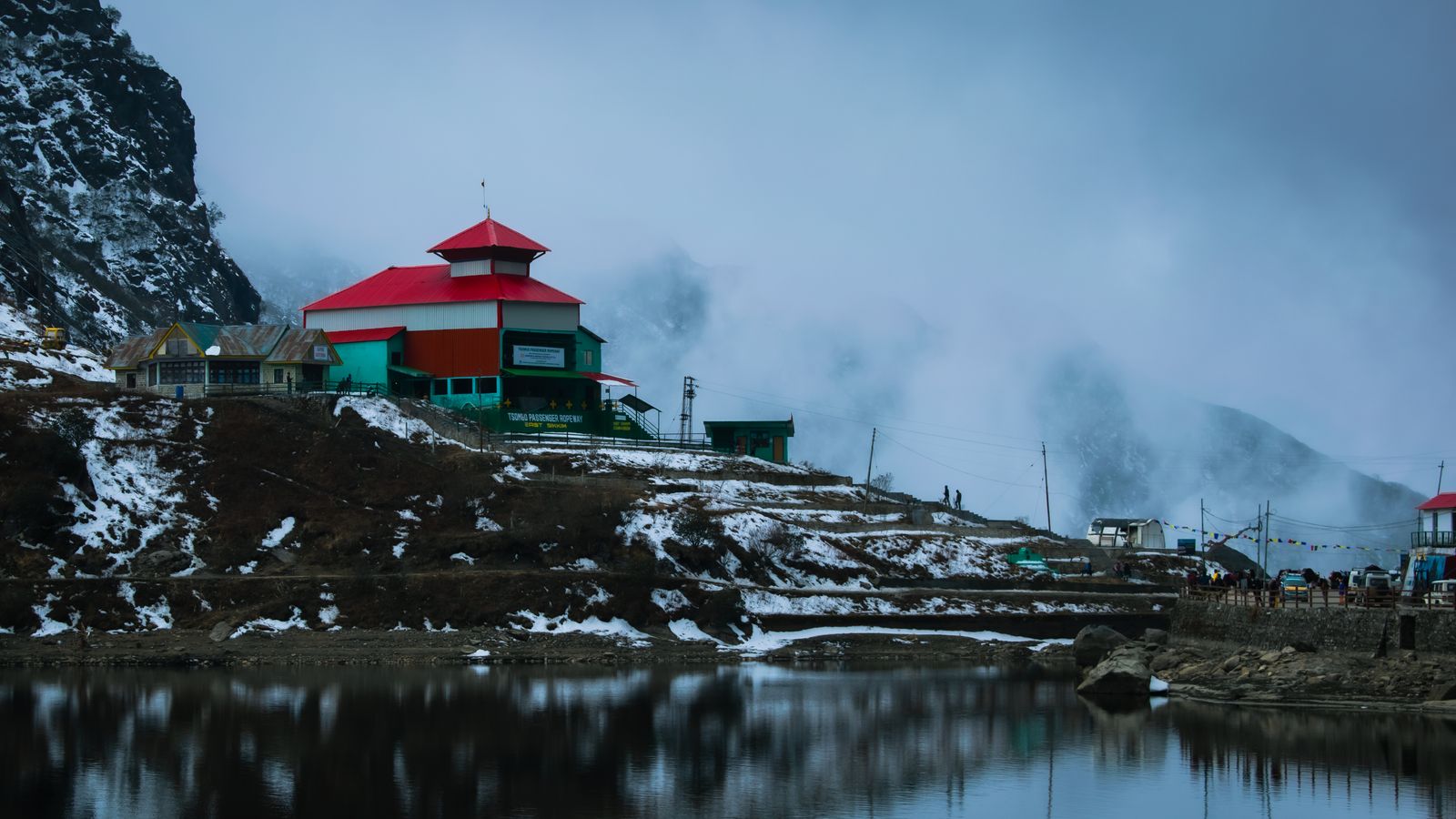 Tsomgo Lake in Sikkim under snow, a red roofed building on the shore and mist rising off the water