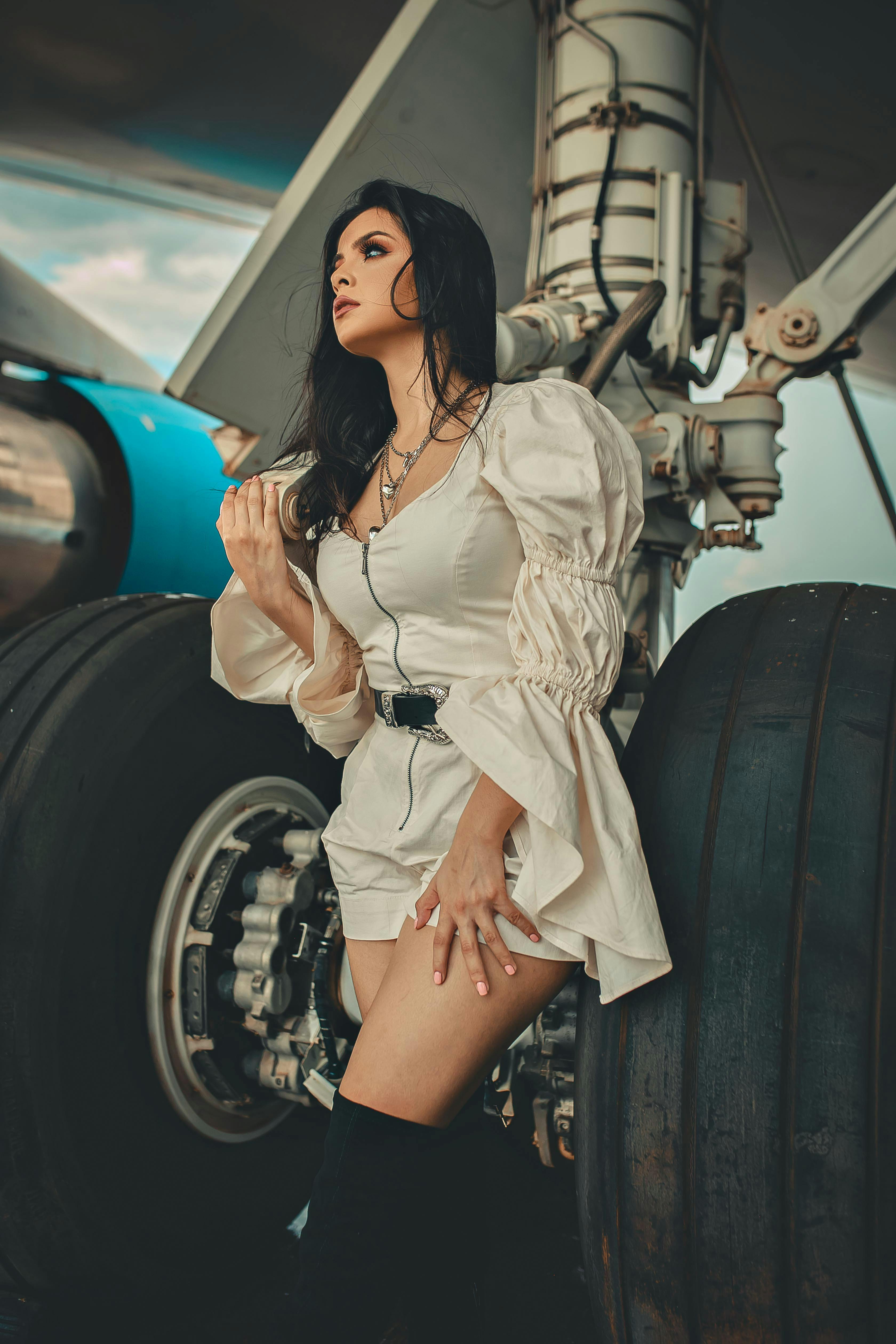 Stylish woman in chic outfit posing dramatically by an airplane's wheel under a clear sky.