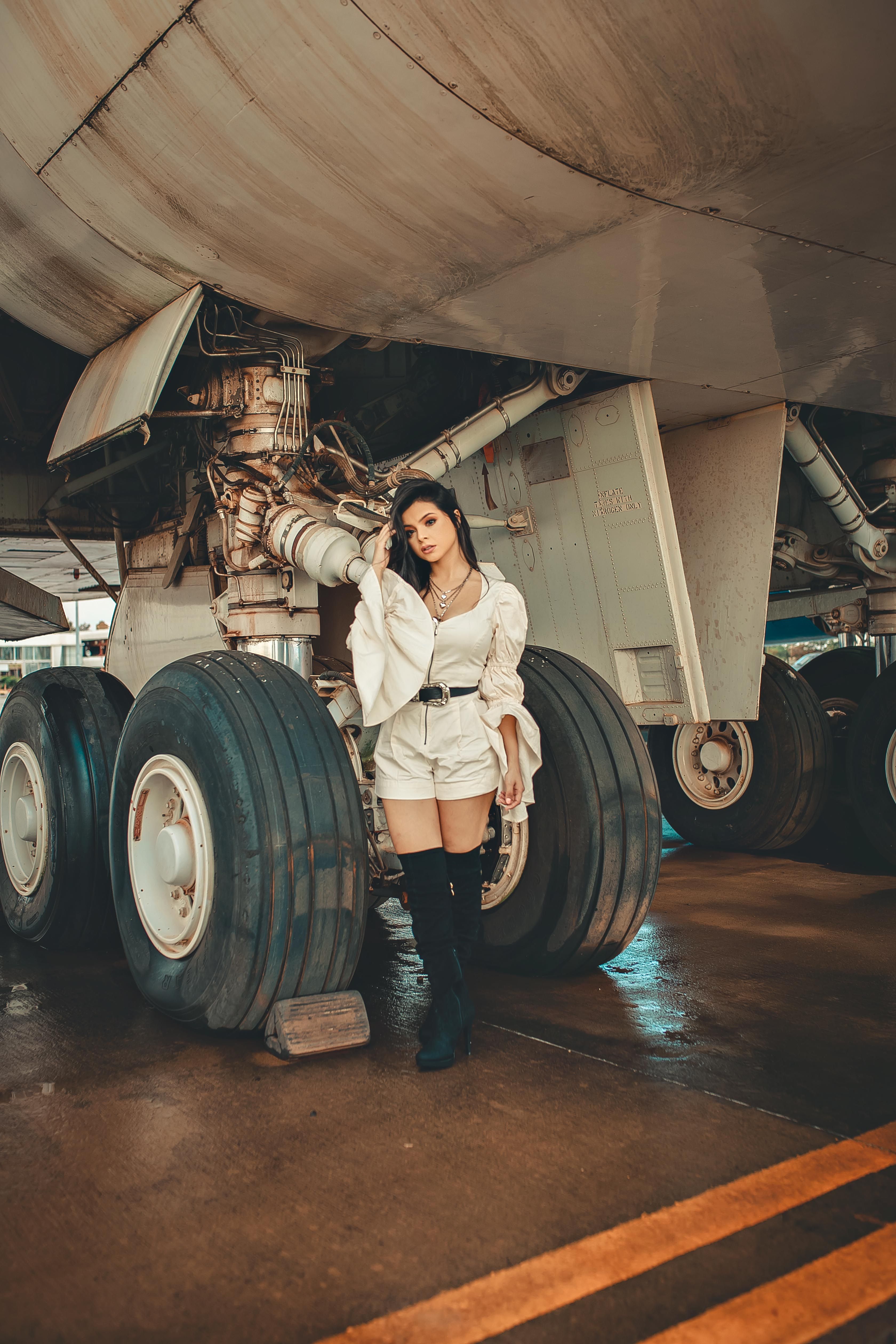 A stylish woman poses under an airplane, showcasing chic fashion in an industrial setting. 