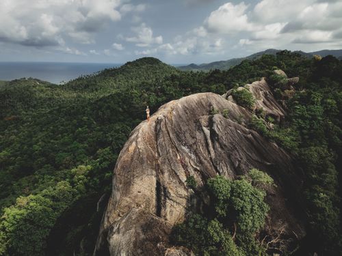 Free Aerial shot of a lone hiker standing on a rugged mountain ridge surrounded by lush green forest under dramatic skies. Stock Photo
