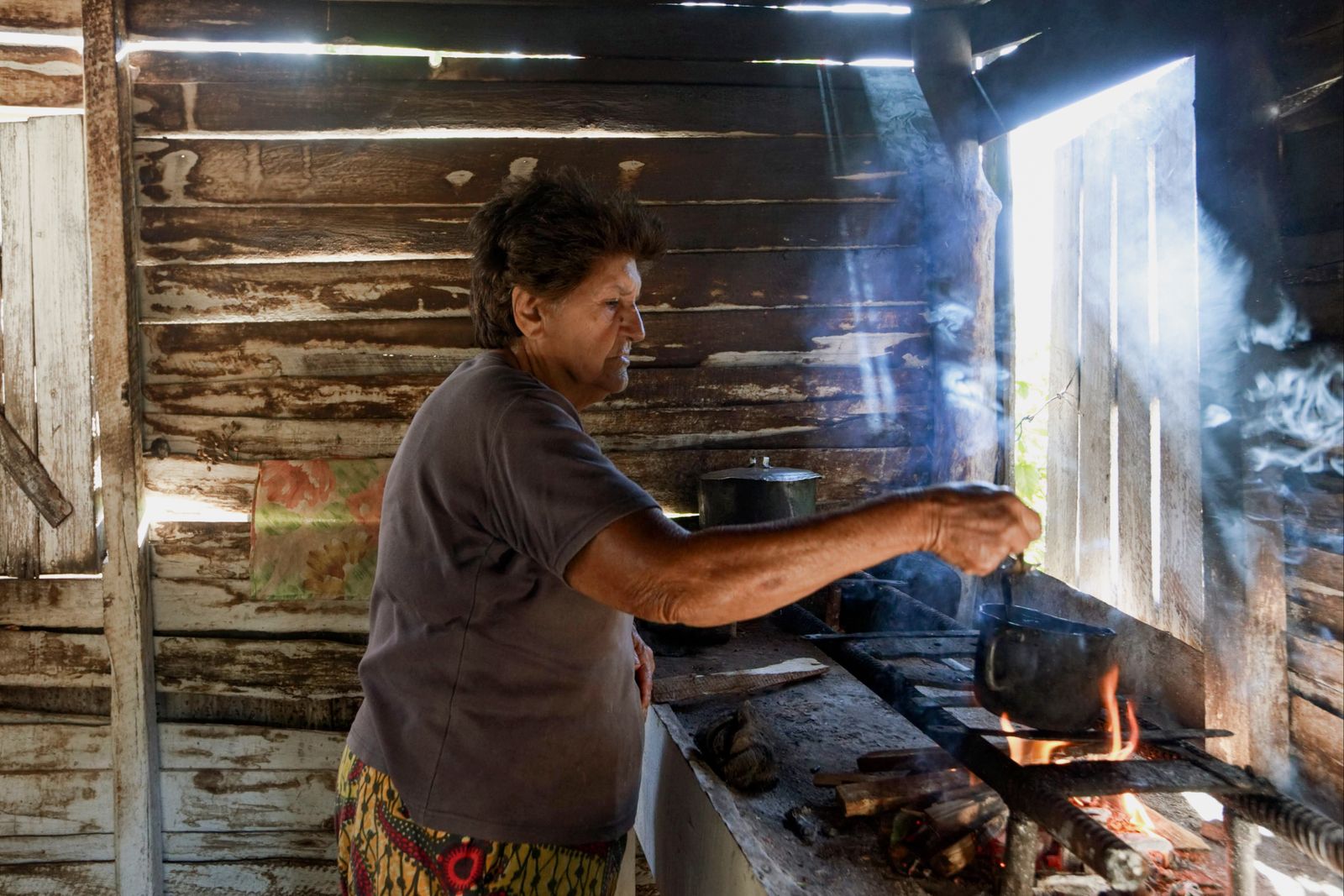 Senior woman cooks over an open flame in a rustic kitchen, evoking tradition and simplicity.