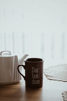 Brown ceramic mug with teapot on a wooden table by lace mat, creating a cozy morning vibe.