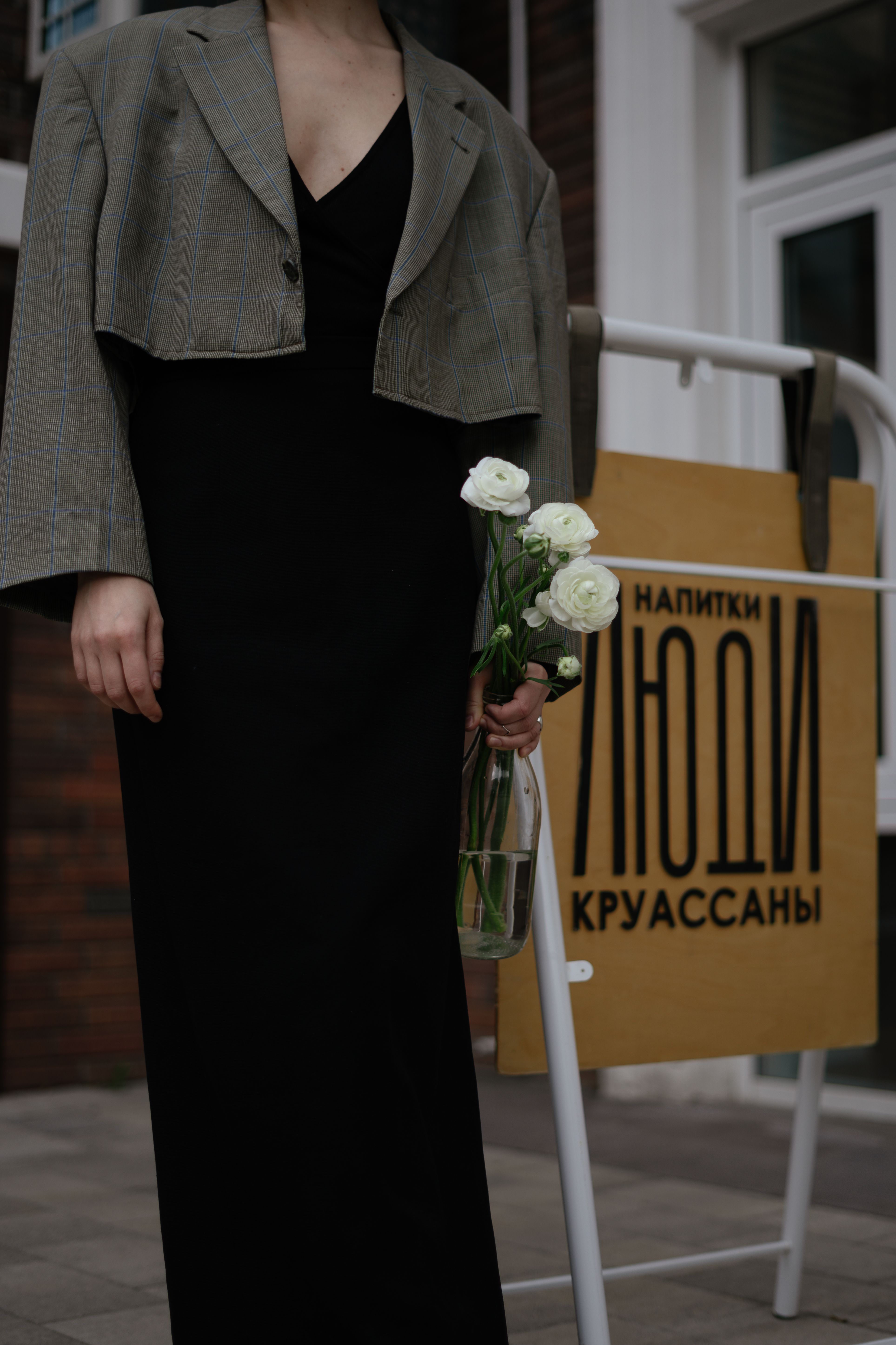 A stylish woman in a black dress holds white roses in front of a cafe sign outdoors.