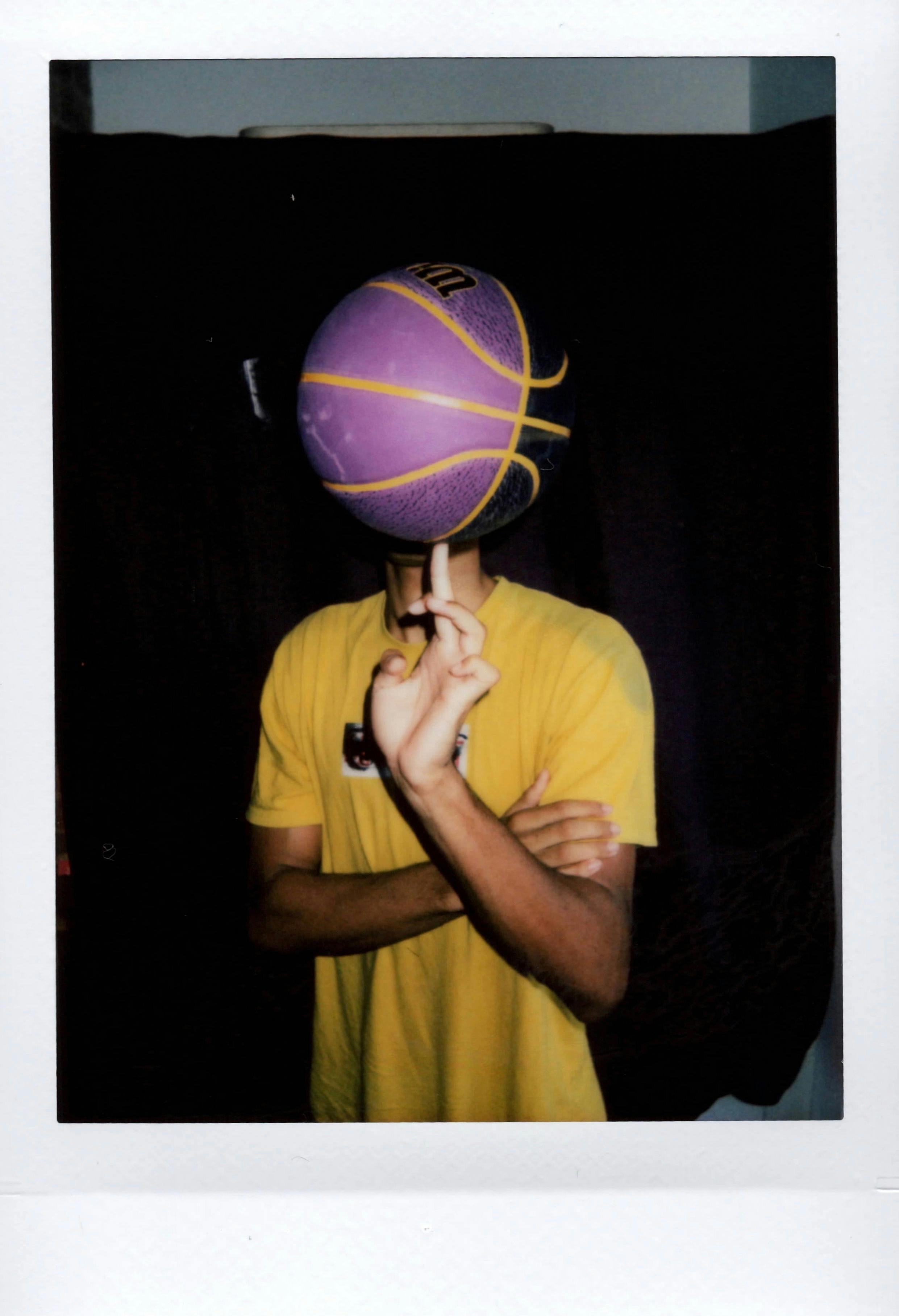 Man in yellow shirt spinning purple basketball on finger indoors.