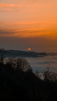 A peaceful sunrise over mist-shrouded hills, with a vibrant orange sky casting a warm glow.