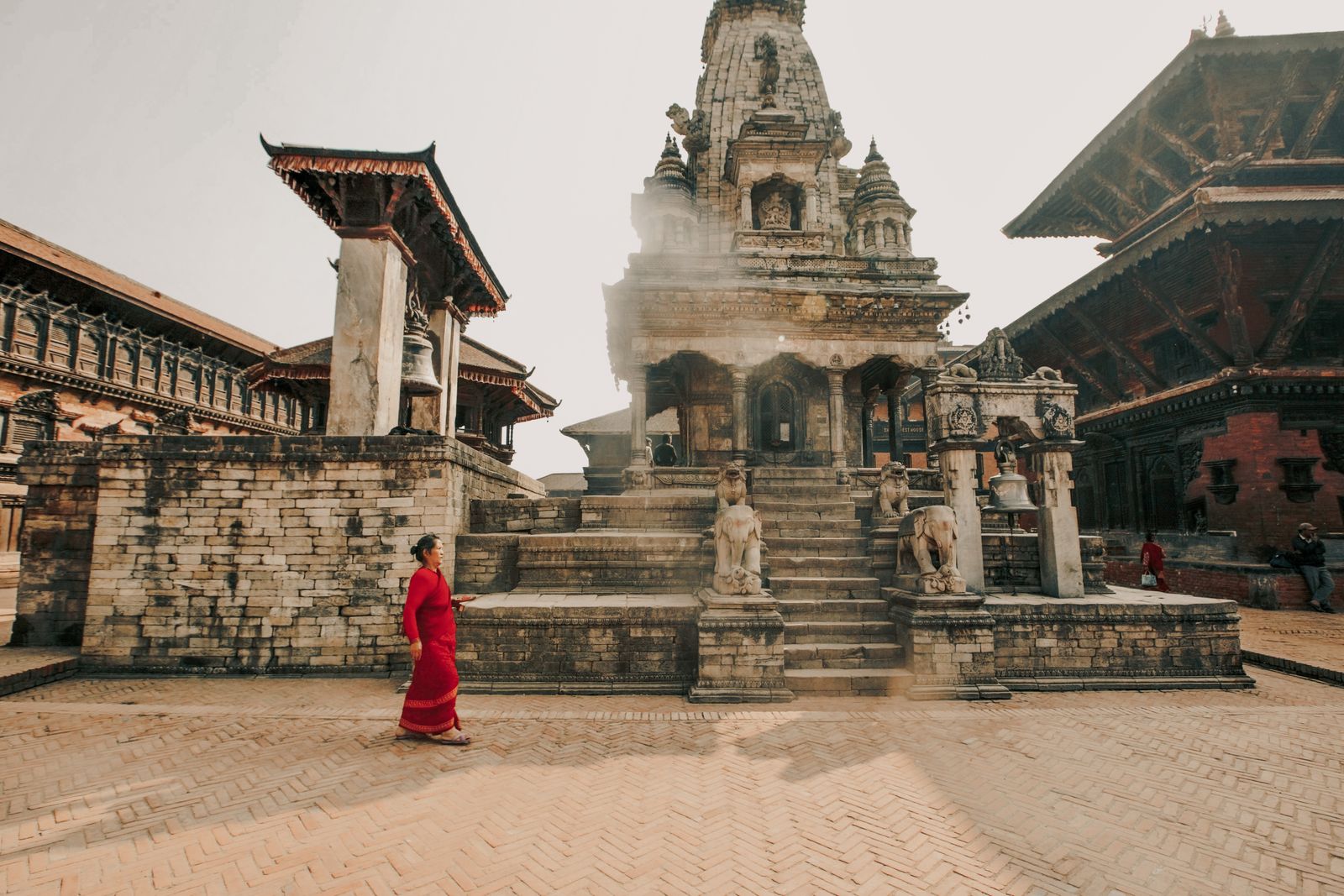 Brick and timber temples around the paved court of Bhaktapur Durbar Square, a woman in red walking past