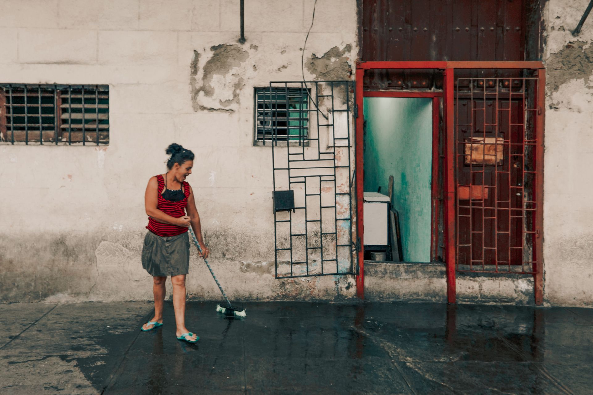 A woman in casual clothing sweeps the pavement outside an aged doorway with red metal bars.