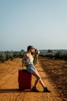 Woman in denim shorts and white tank top resting by luggage on a sunny dirt pathway, looking into the distance.