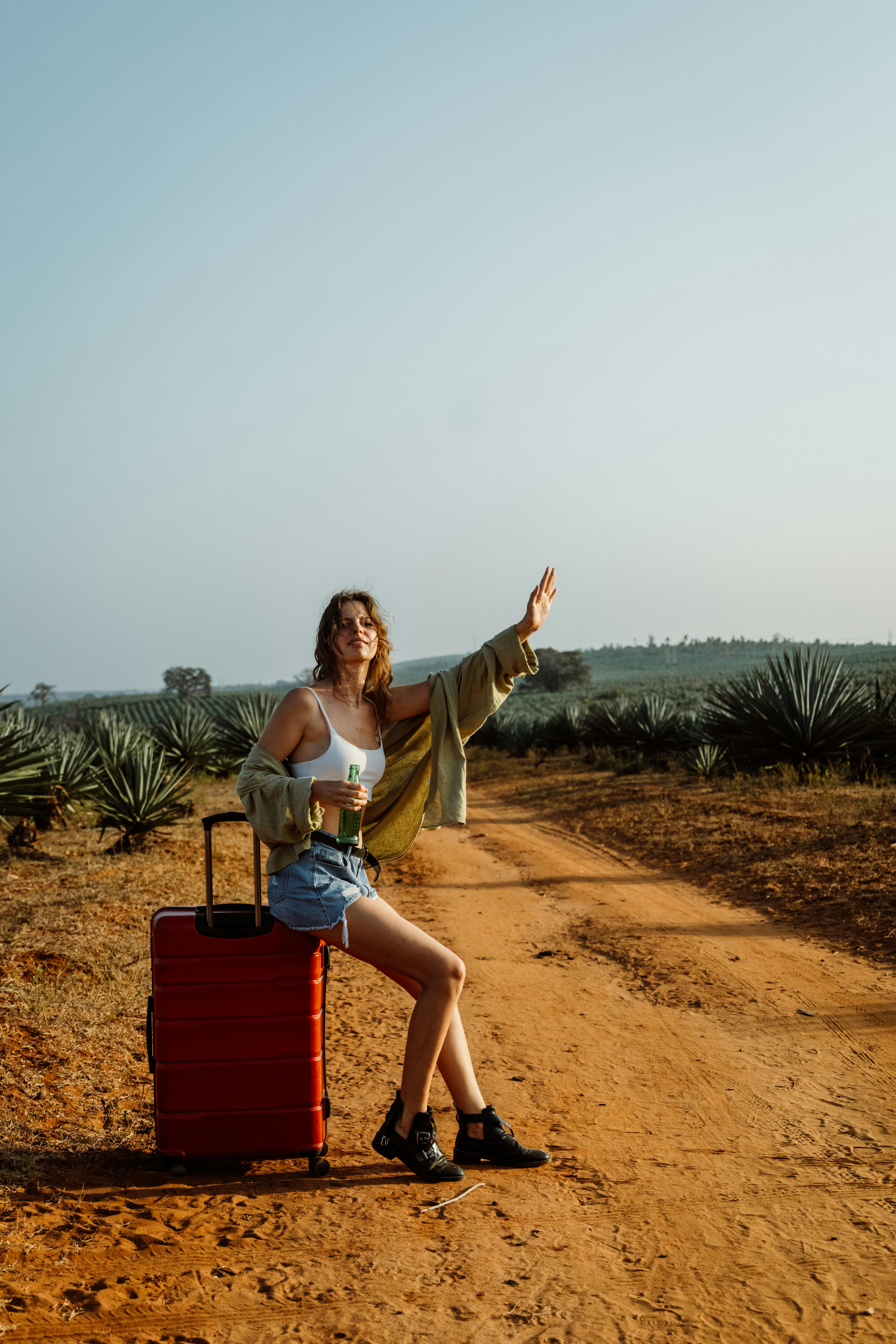 Fashionable woman on a dirt road with a red suitcase, hitchhiking in a desert landscape.