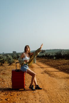 Fashionable woman on a dirt road with a red suitcase, hitchhiking in a desert landscape.