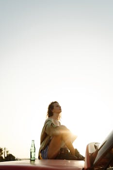 Young woman sitting on a car under bright sunlight, enjoying a peaceful moment outdoors.