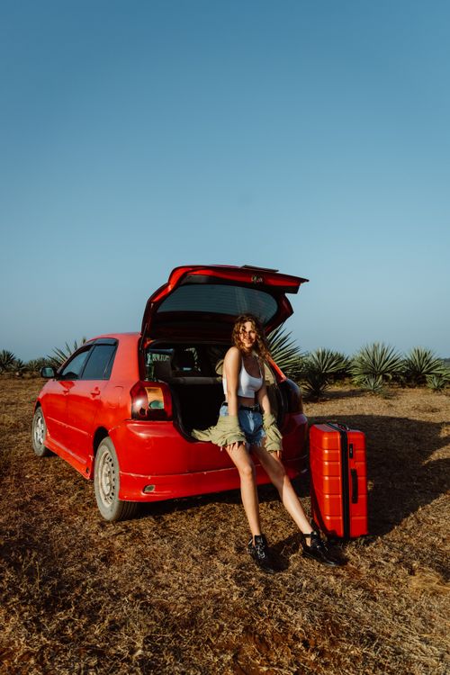 Free Young woman in denim shorts posing with suitcase beside a red car in a field. Stock Photo