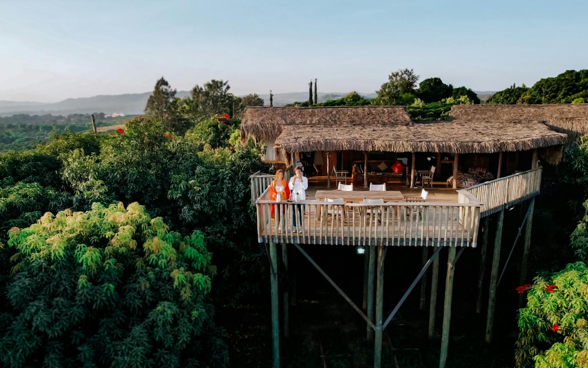 Aerial view of a couple relaxing on a treehouse deck in a lush tropical setting.