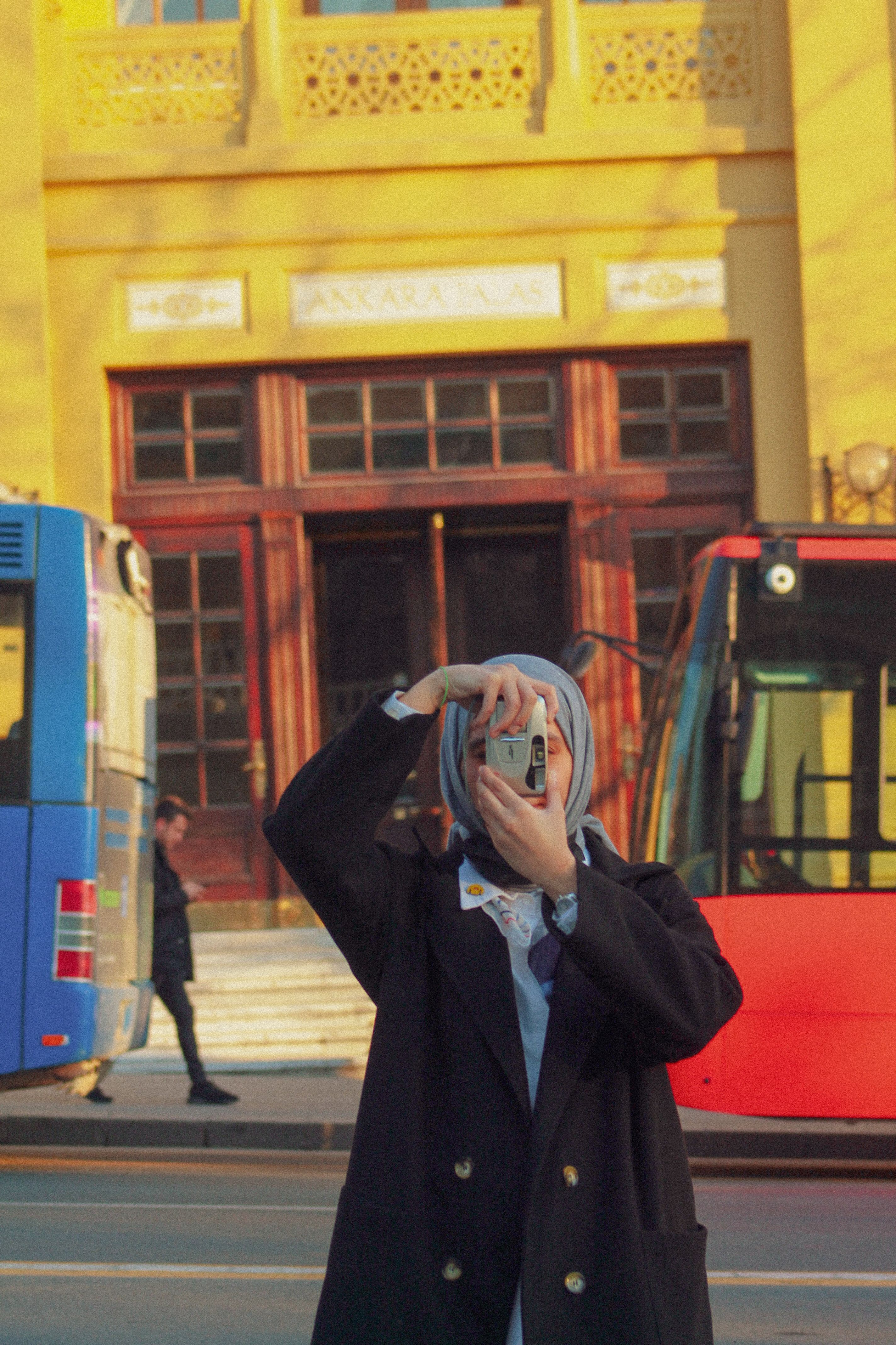 A woman in a headscarf using an analog camera to capture a city scene, standing between colorful buses.