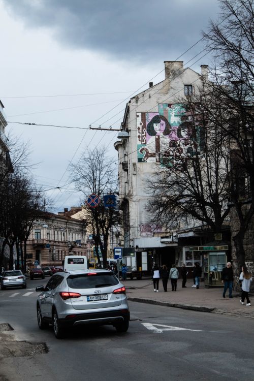 Free City street with mural-filled buildings, cars, and pedestrians on a cloudy day. Stock Photo