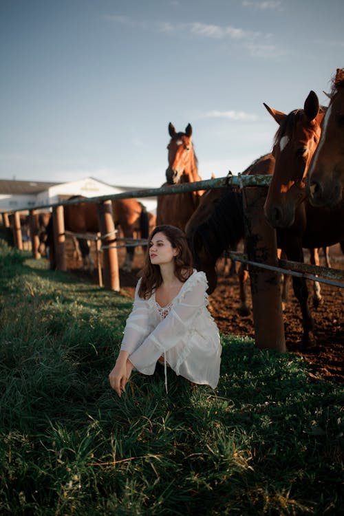 Woman in White Dress with Horses in Pen Behind 