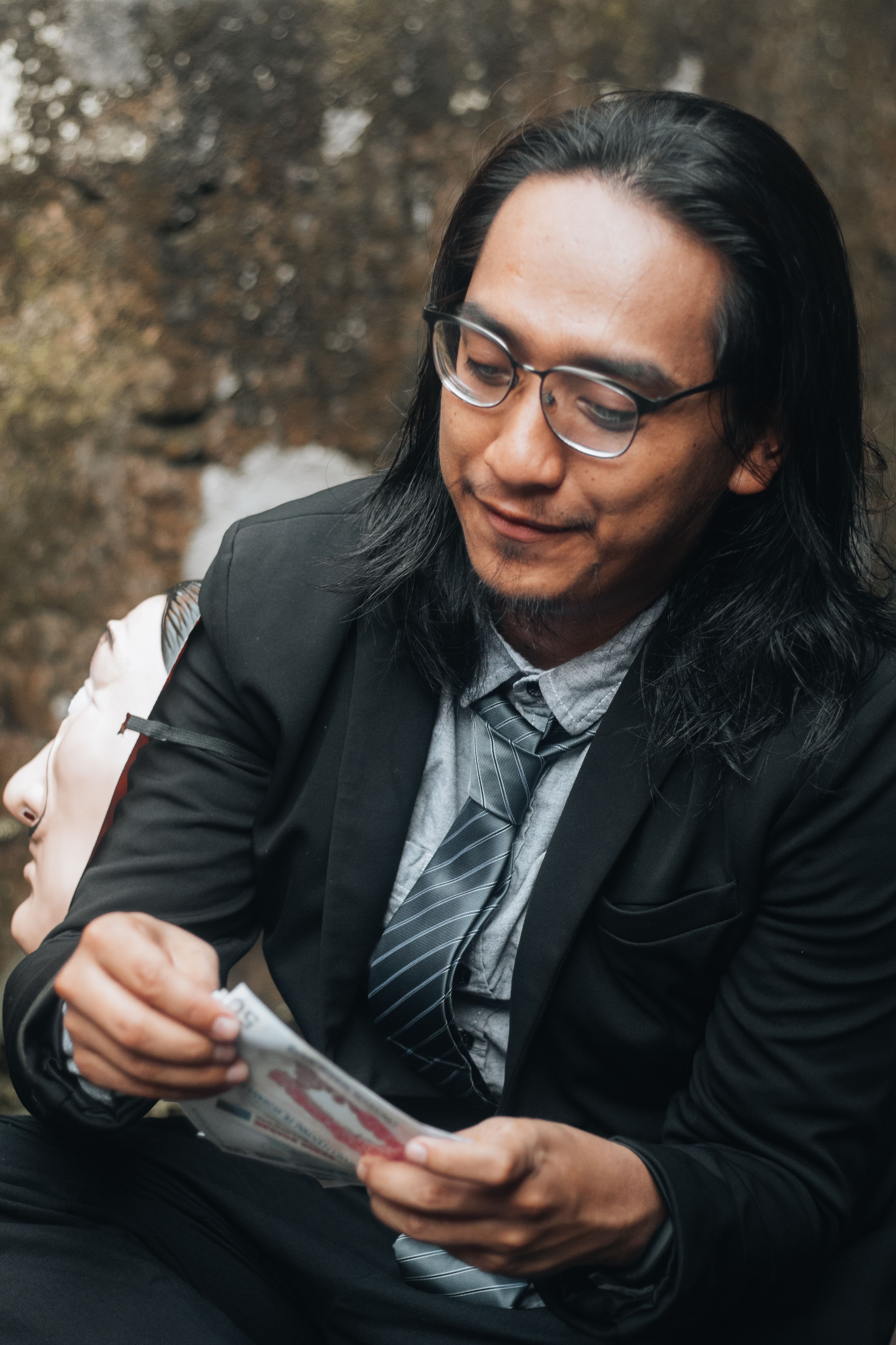 Asian man in business attire holding a mask in a contemplative moment outdoors.