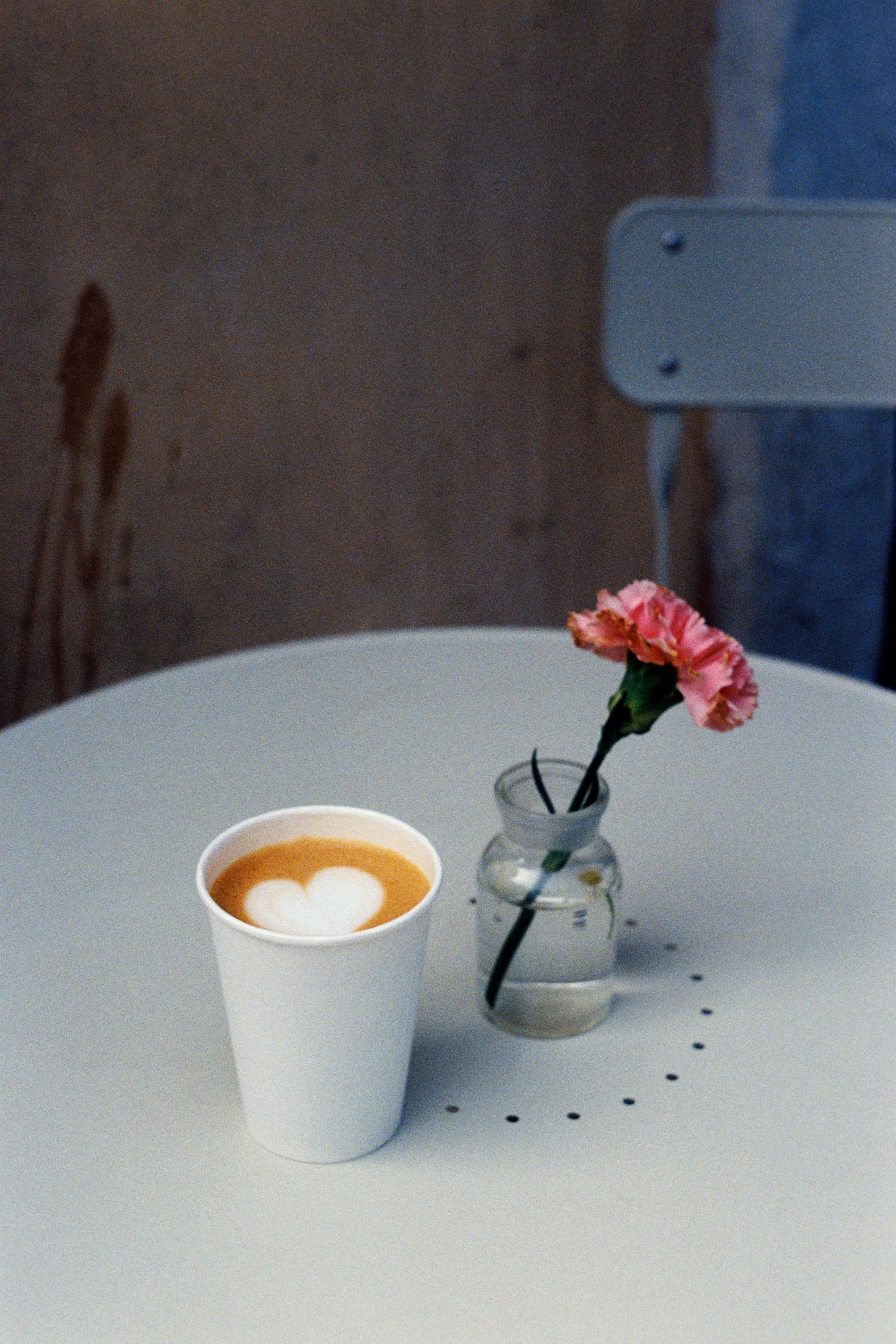 A white cup of latte with heart art beside a pink flower in a vase on a white table.