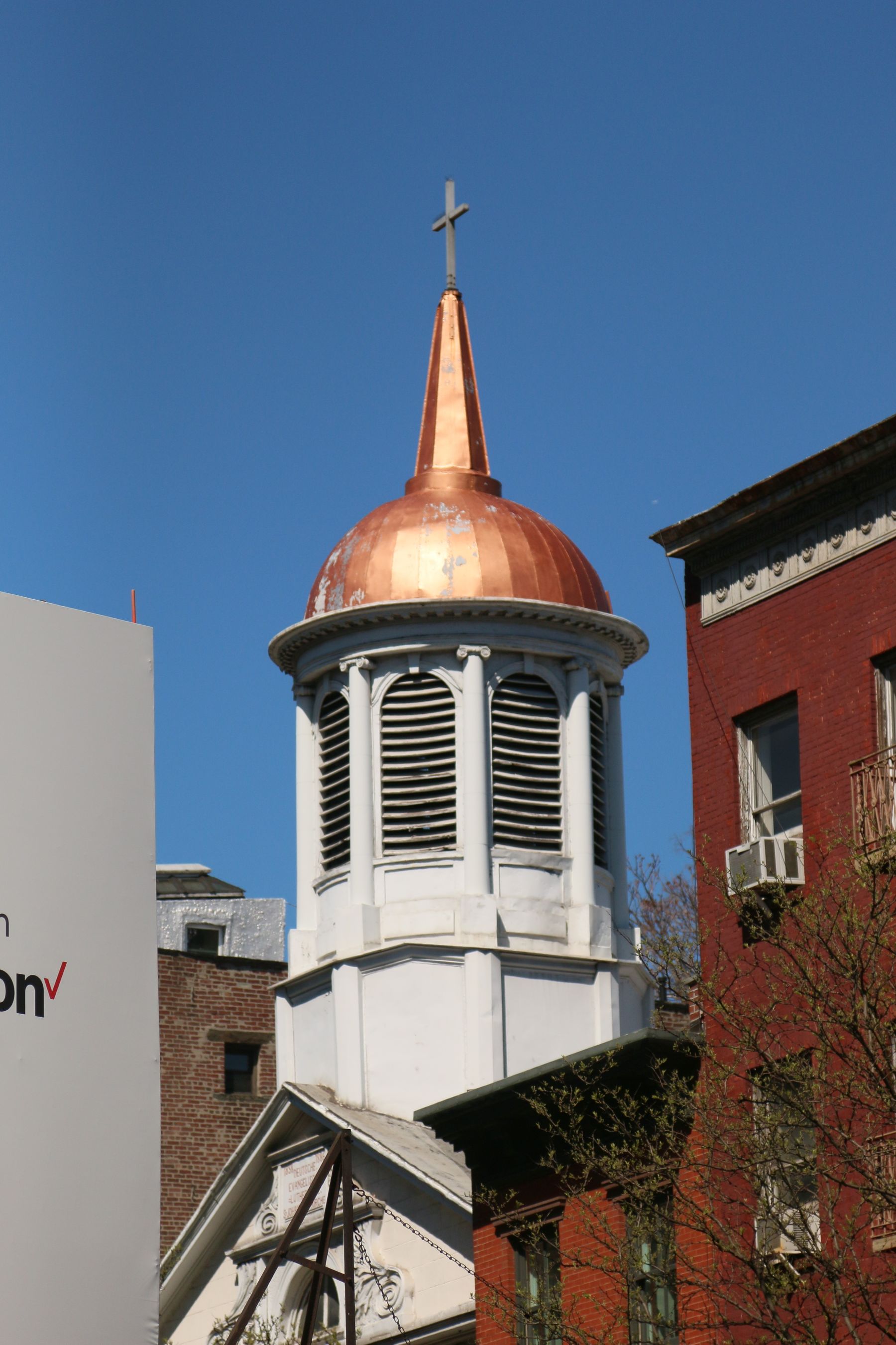 Copper-colored dome and cross atop a louvered tower between buildings under a blue sky.