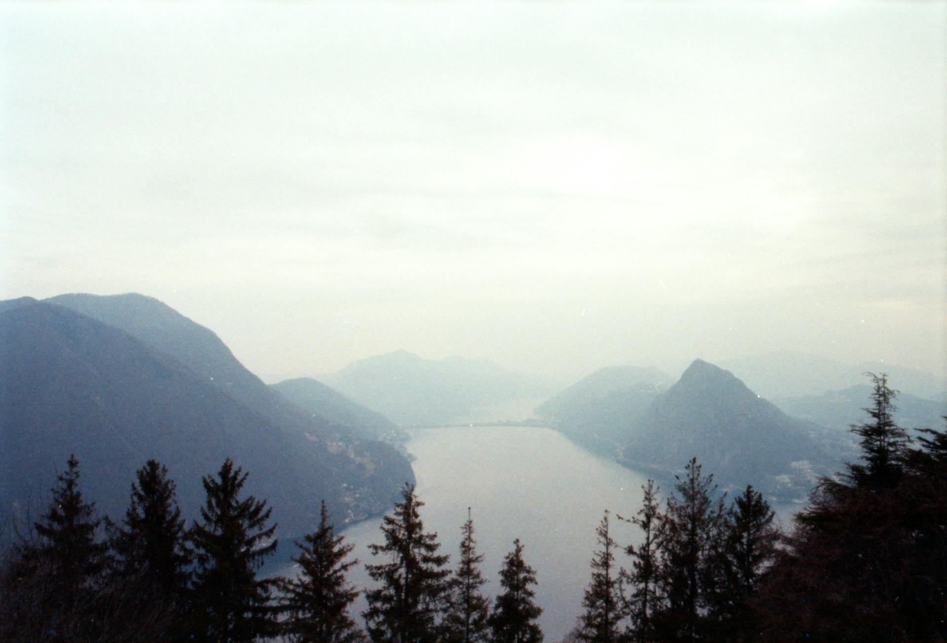 A serene misty view of Lake Lugano with surrounding mountains and conifer trees.