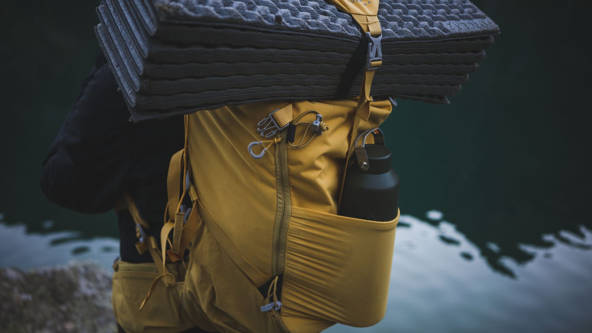 Yellow backpack with camping gear including a water bottle and folded mat near water.