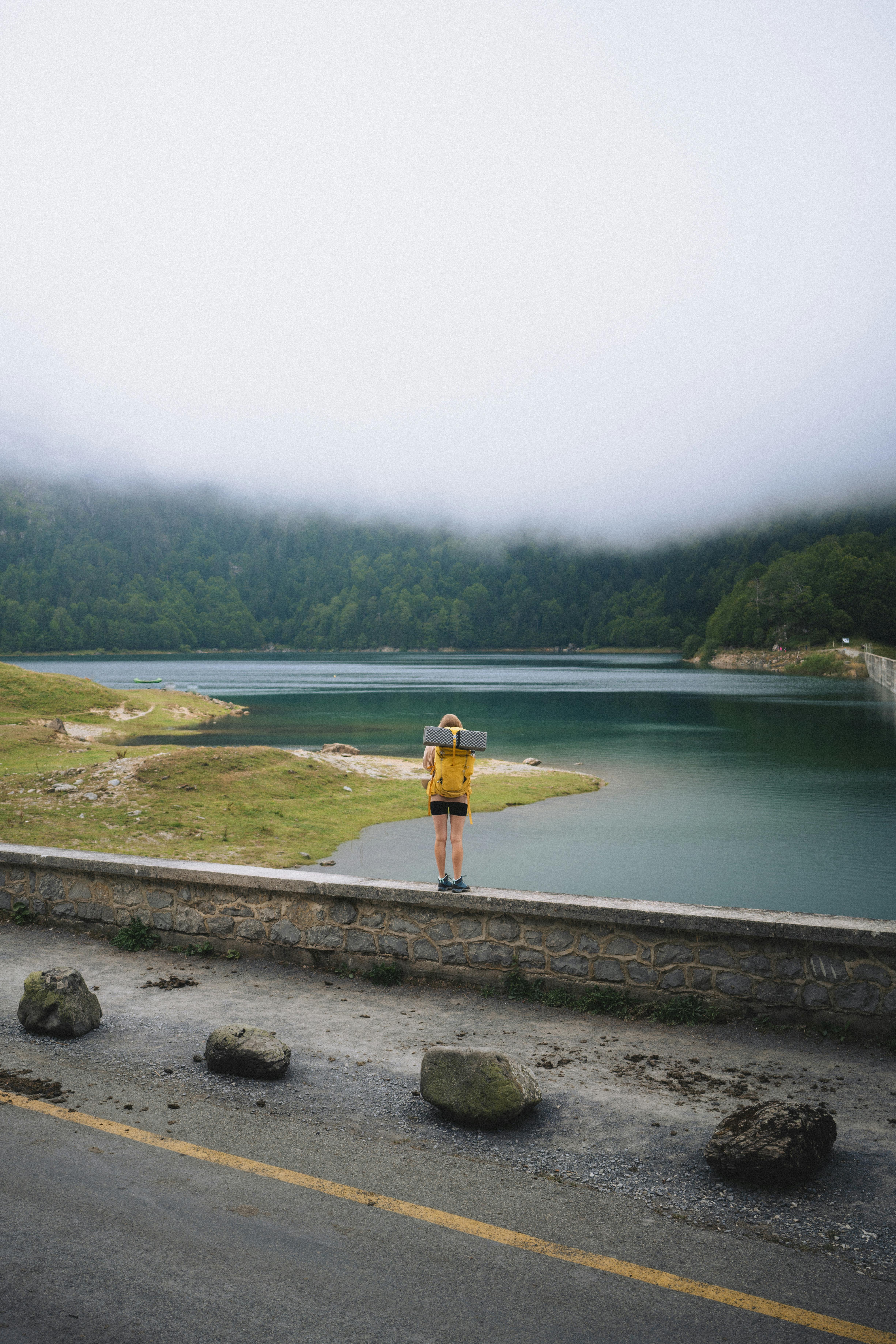 A lone backpacker stands by a fog-covered lake, surrounded by mountains, embracing adventure.