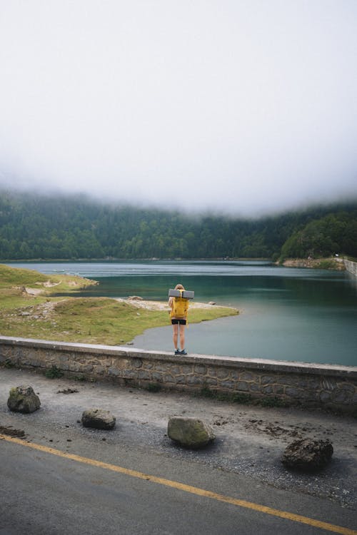 Woman Standing on the Verge of a Road Beside Lake in Mountains 