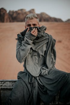 Man in gray robe sitting in desert, hand covering face. Moody and mysterious vibe.