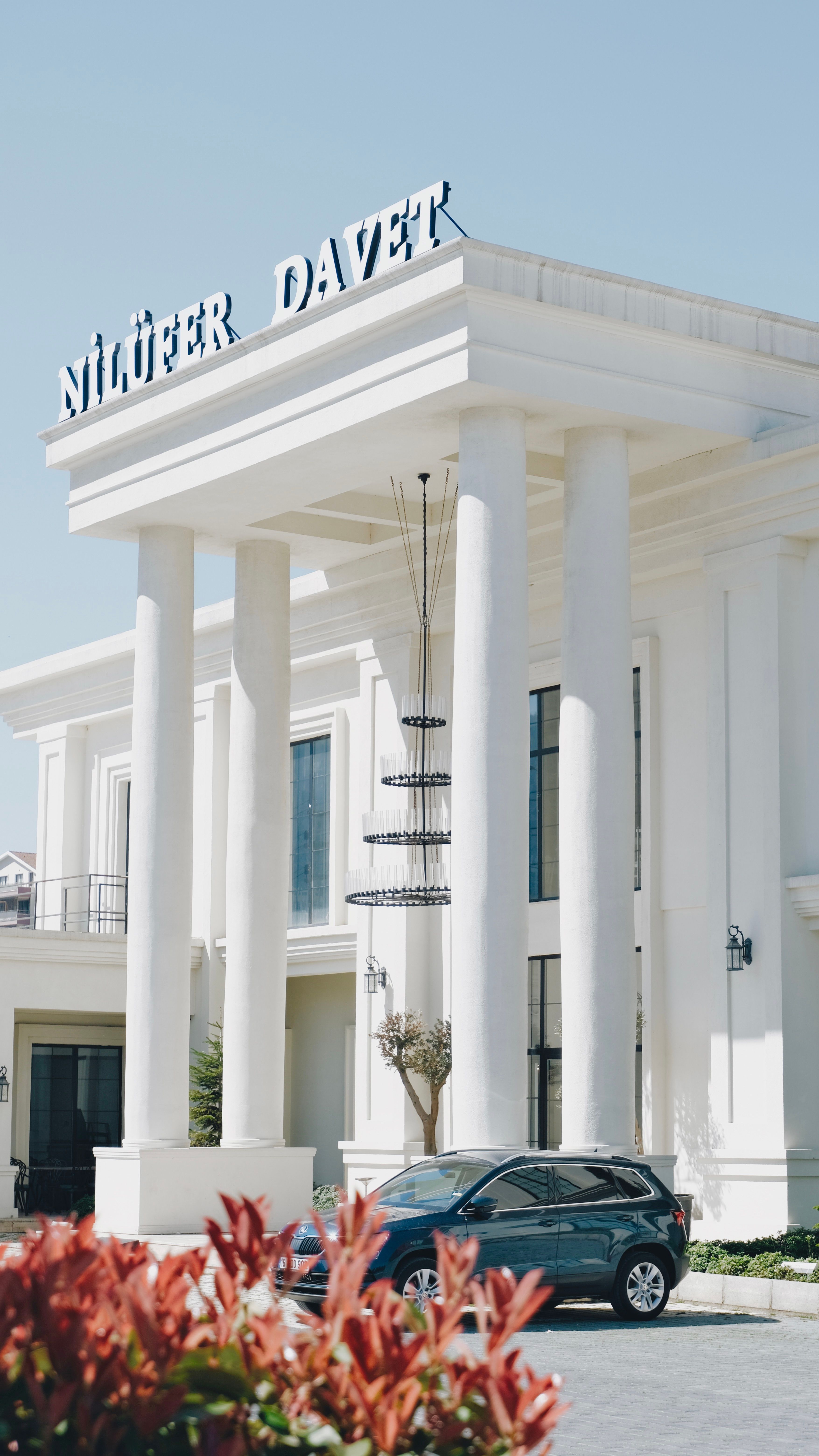 A modern white building with grand columns and a parked car in front, viewed during the day.