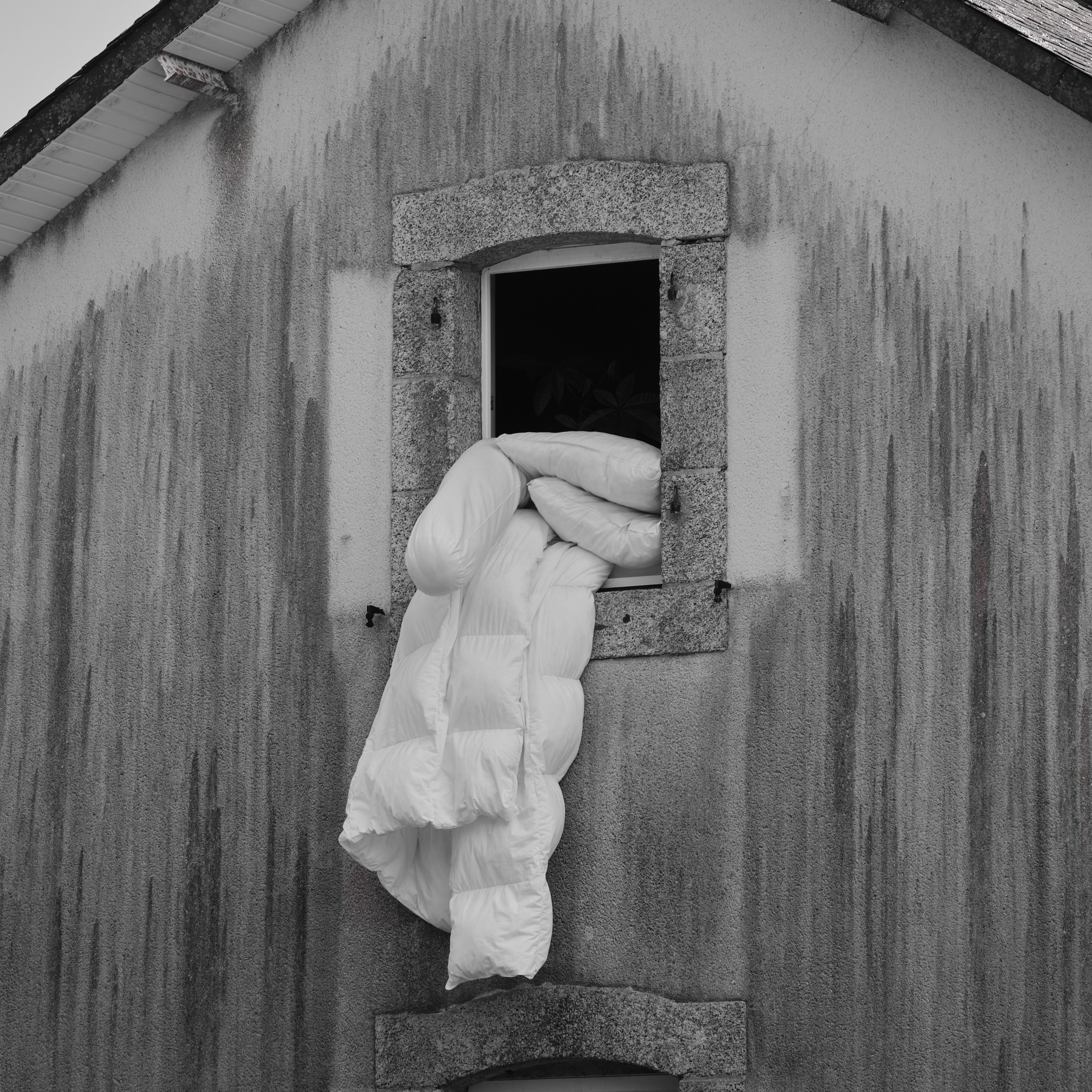 A monochrome image of a duvet draped over a rustic building's windowsill.