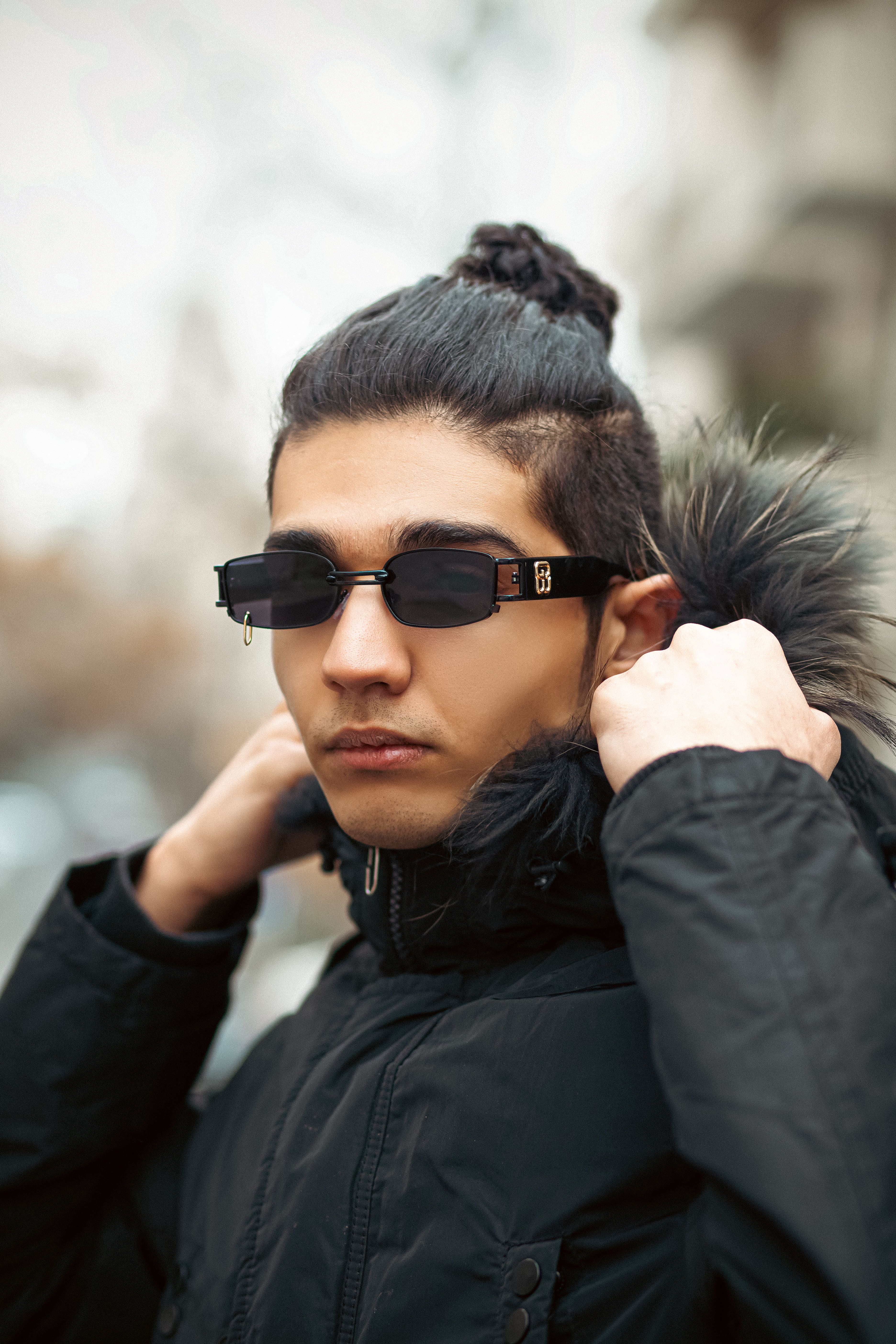Portrait of a fashionable man with sunglasses adjusting a black jacket outdoors.
