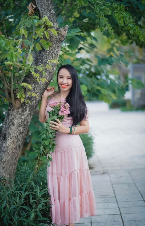 Free Woman in a pink dress standing outdoors, holding flowers with a bright smile. Stock Photo