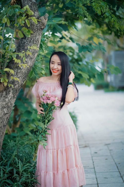 Free Beautiful woman in a pink dress holding flowers, posing outdoors surrounded by lush greenery. Stock Photo
