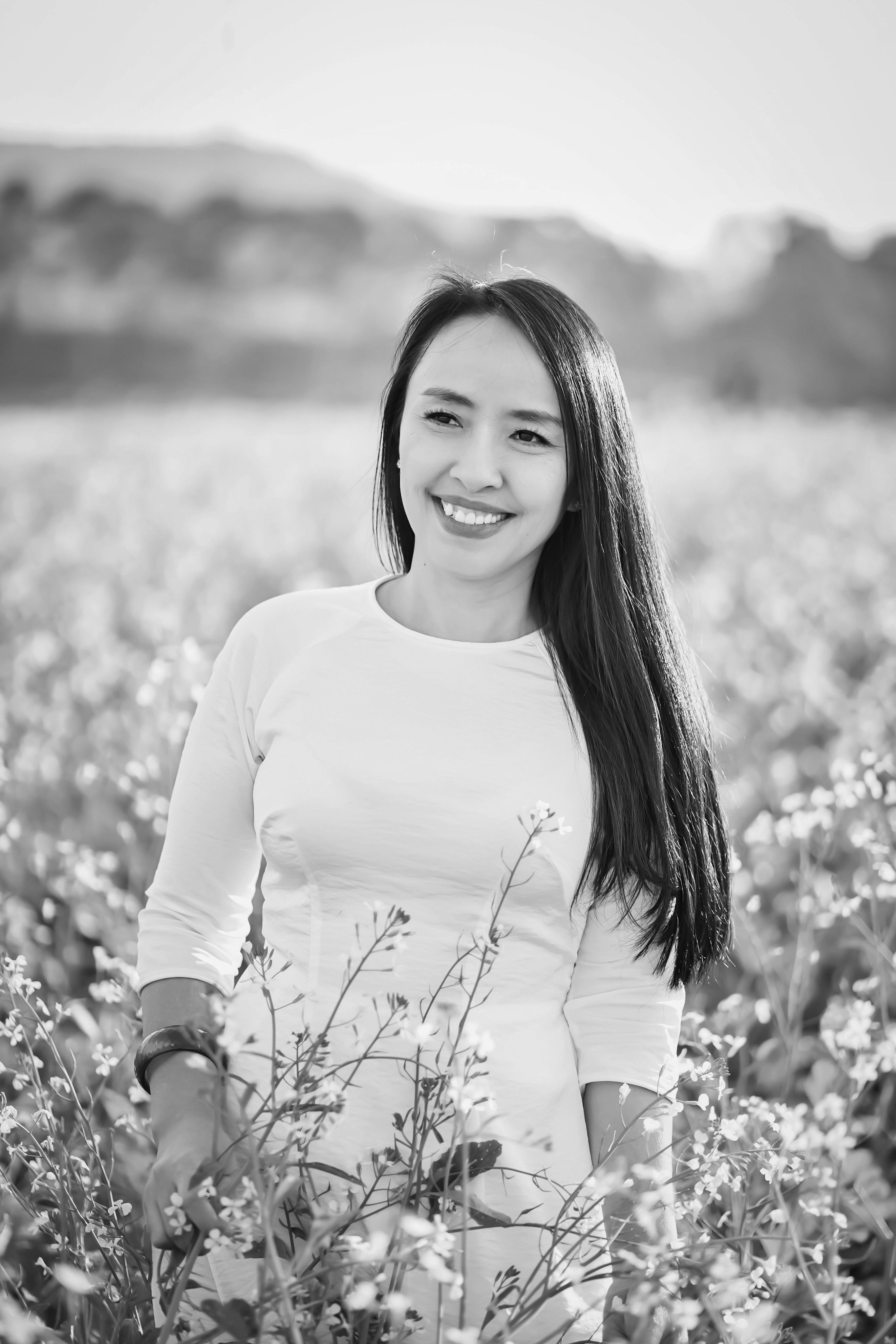 A woman with long hair smiling in a black and white field setting, surrounded by flowers.