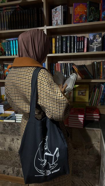 A woman in a hijab reads a book while standing in front of a bookshelf in a library. - best handbag for personal style in Pakistan