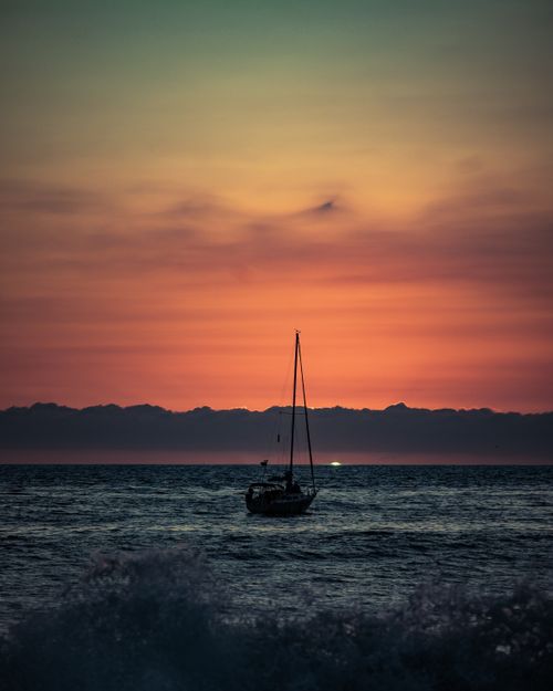 Free A lone sailboat silhouetted against a vibrant orange sunset over the calm sea waves. Stock Photo