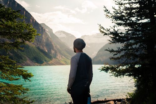 Free A man in casual attire stands by a picturesque mountain lake, surrounded by lush greenery. Stock Photo