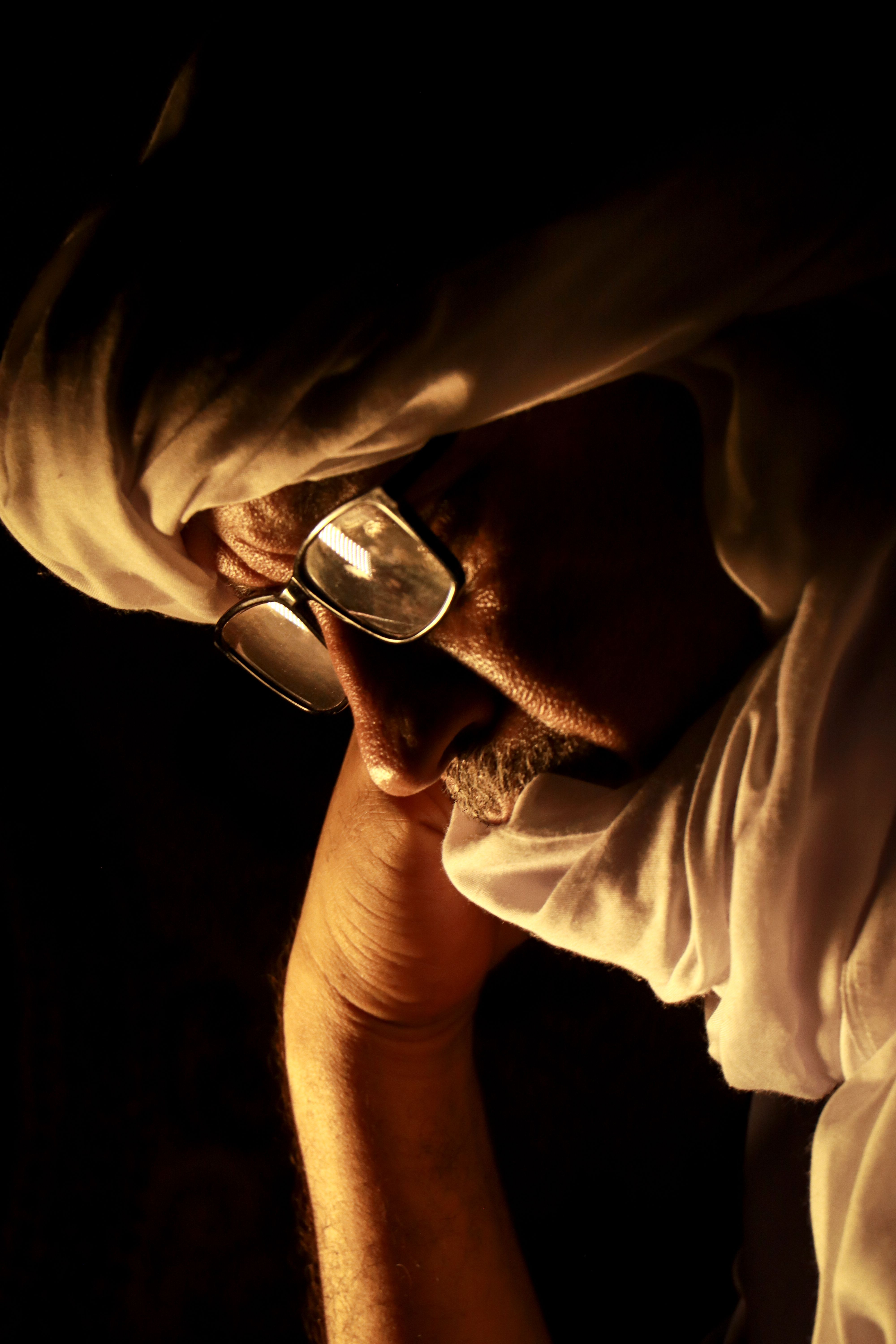 A thoughtful man wearing traditional attire and glasses in dim lighting, embodying introspection.
